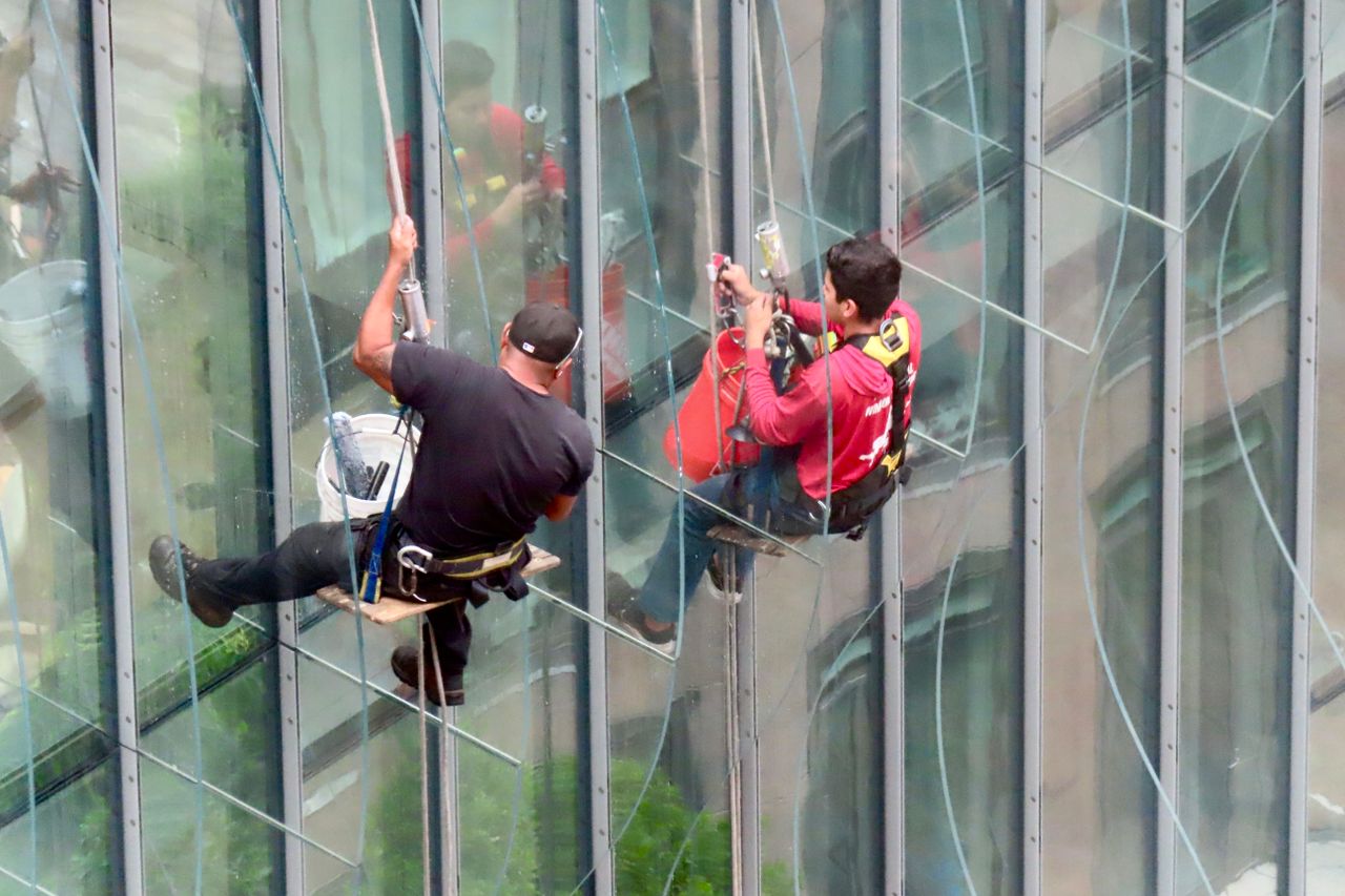 services-03 Deux nettoyeurs de vitres professionnels à l’œuvre sur un immeuble vitré de grande hauteur, équipés de harnais suspendus.